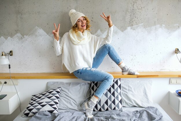 Beautiful young woman sitting in bedroom against wall wearing white sweater