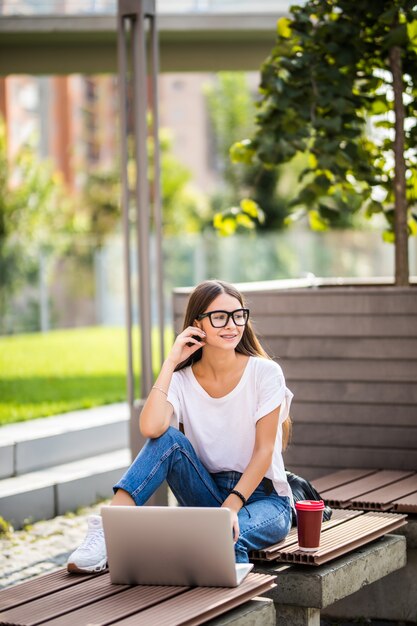 屋外のラップトップを使用しながらコーヒーを保持しているベンチに座っている美しい若い女性。