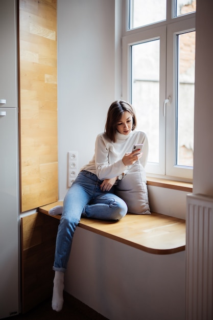 Beautiful young woman sitting on wide windowhill in blue jeans and white t-shirt