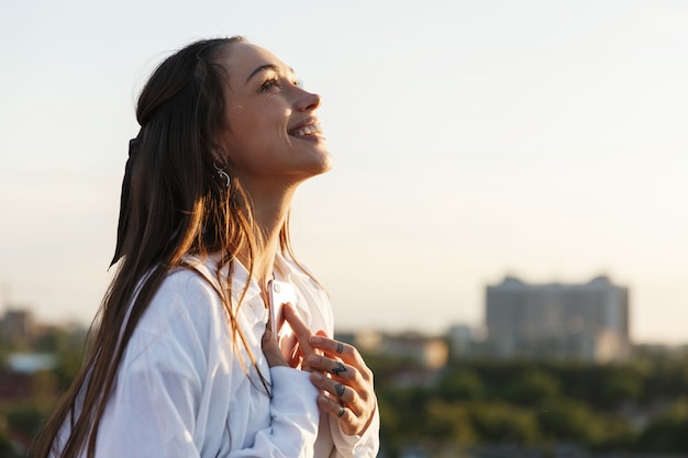 Beautiful young woman smiles tender standing on the rooftop in the rays of evening sun