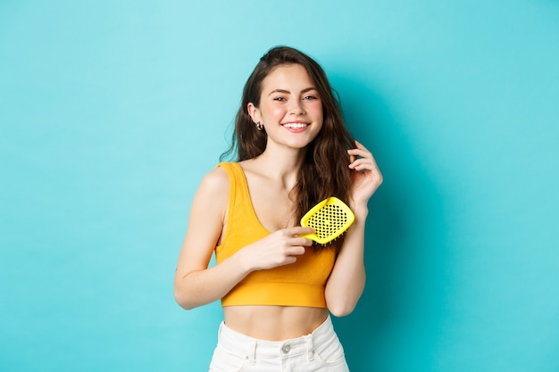 Beautiful young woman smiling, brushing healthy long hair with hairbrush, standing against blue background