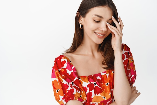 Beautiful young woman smiling coquettish touching her face and looking down shy blushing standing in floral dress against white background