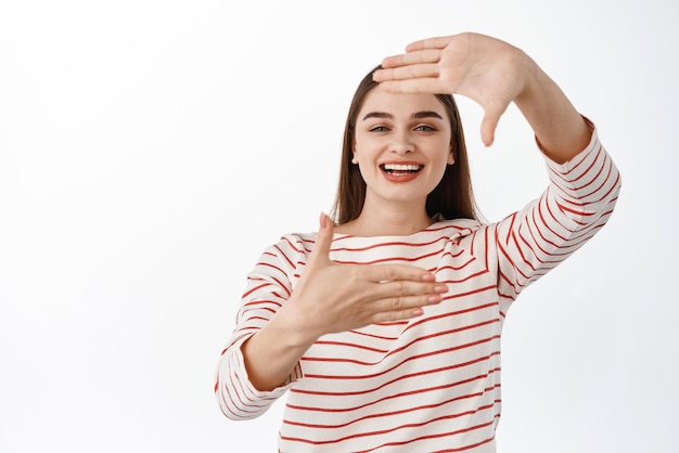 Beautiful young woman smiling looking through hand frames as if imaging searching perfect angle taking photo in her mind standing over white background