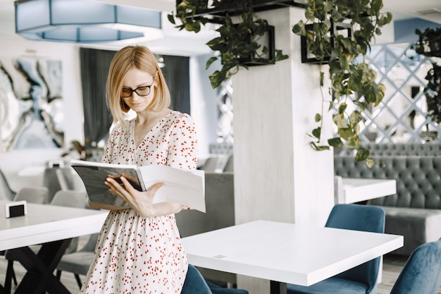 A beautiful young woman standing in a cafe indoors and reading some documents. Woman wearing glasses and floral dress