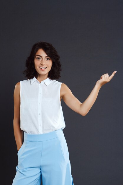 Beautiful young woman standing over grey wall and pointing her finger