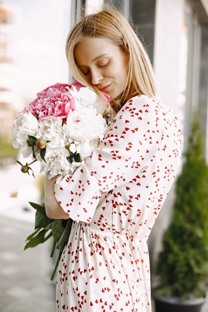 屋外のカフェの近くに立って、花の花束を持っている美しい若い女性。花柄のドレスを着ている女性