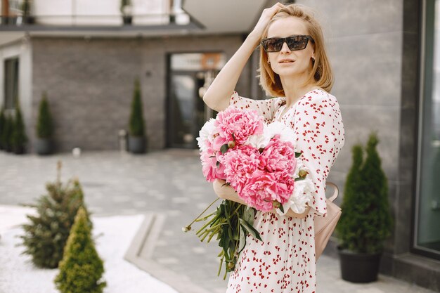 屋外のカフェの近くに立って、花の花束を持っている美しい若い女性。眼鏡とドレスを着ている女性