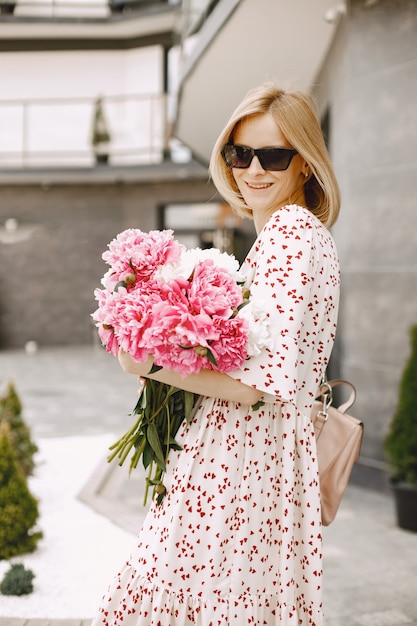 屋外のカフェの近くに立って、花の花束を持っている美しい若い女性。眼鏡とドレスを着ている女性