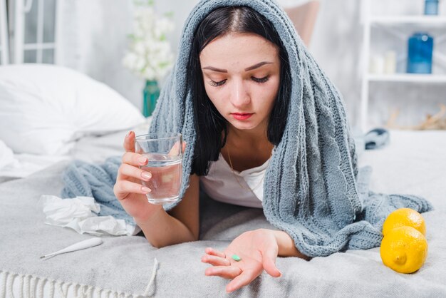 Красивая молодая женщина страдает от лихорадки, держа в руке стакан воды и таблетки