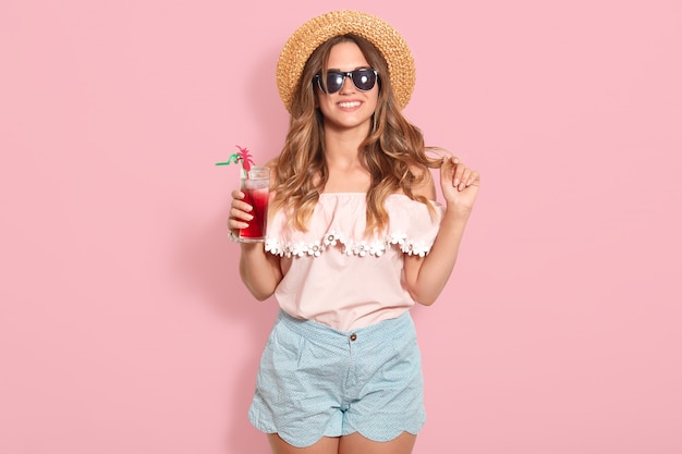 Beautiful young woman in summer blouse, blue short, black sunglasses and straw hat holding glass