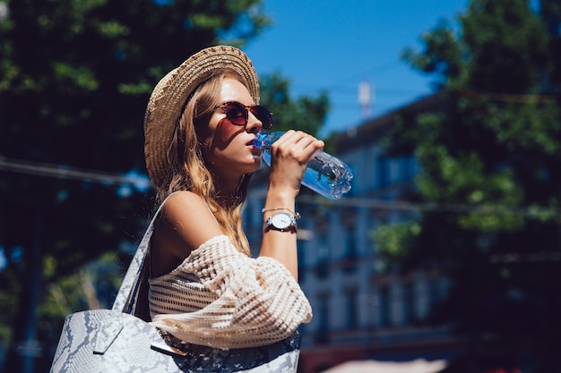 Beautiful young woman in sunglasses, drinking a water from bottle, walking outdoors.