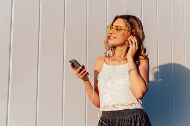 彼女の電話でイヤホンで音楽を聴くサングラスの美しい若い女性