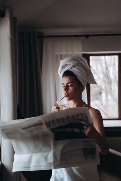 Beautiful young woman in a towel smokes a cigarette and reads newspaper