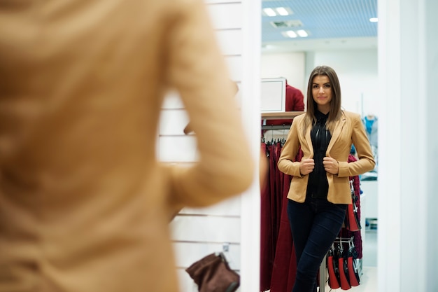 Beautiful young woman trying on jacket in front of mirror