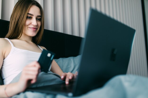 Красивая молодая женщина, используя свою кредитную карту в Интернете, делая покупки, лежа на кровати