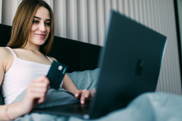 Beautiful young woman using her credit card on the web shopping lying on the bed