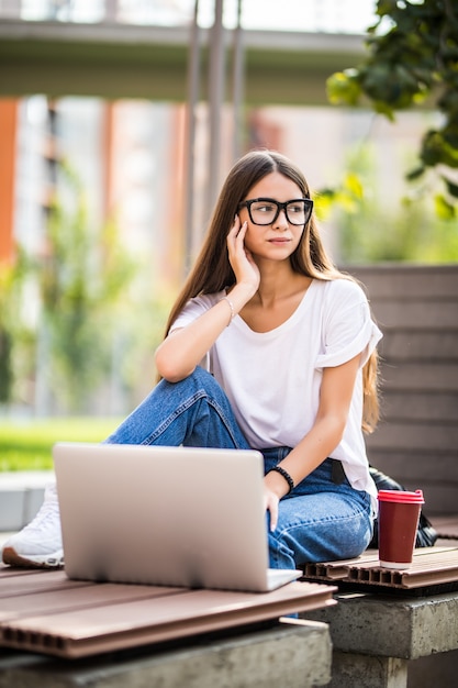 Foto gratuita bella giovane donna che per mezzo del computer portatile mentre sedendosi su un banco, bevendo la tazza di caffè asportabile