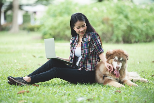 Beautiful young woman using laptop with her little dog in a park outdoors. Lifestyle portrait.