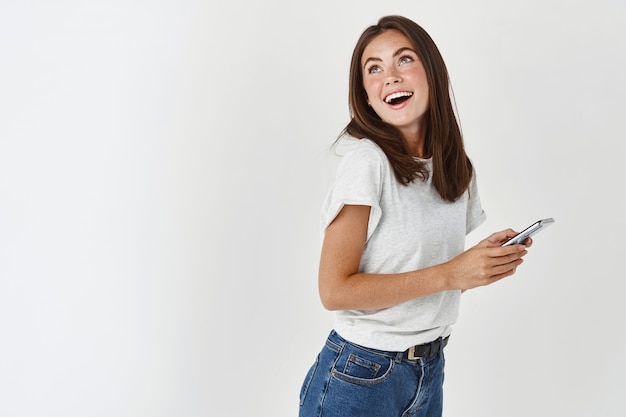 Beautiful young woman using smartphone, smiling and dreamy looking at upper left corner logo, standing over white wall.