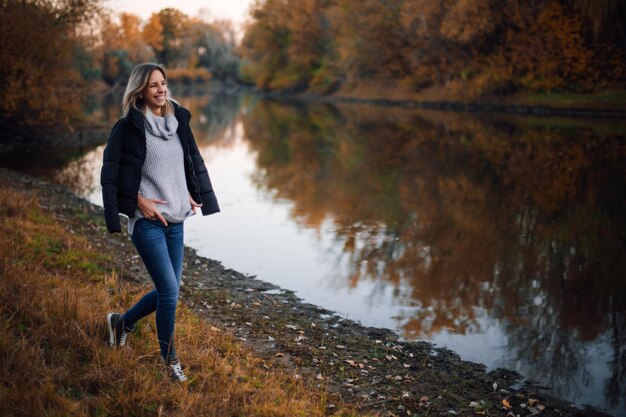 Beautiful young woman walking towards river looking away smiling with teeth with black jacket on sho...