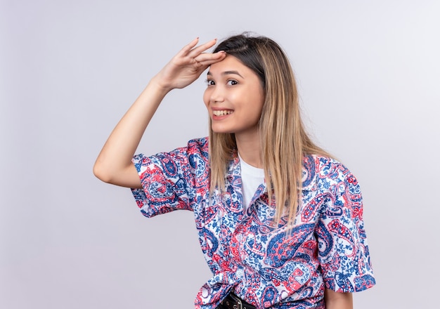 A beautiful young woman wearing paisley printed shirt looking far away and searching for something on a white wall