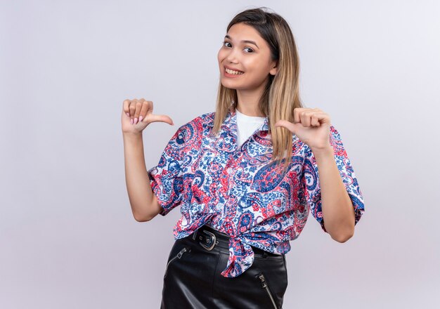 A beautiful young woman wearing paisley printed shirt pointing thumb finger at herself while looking on a white wall