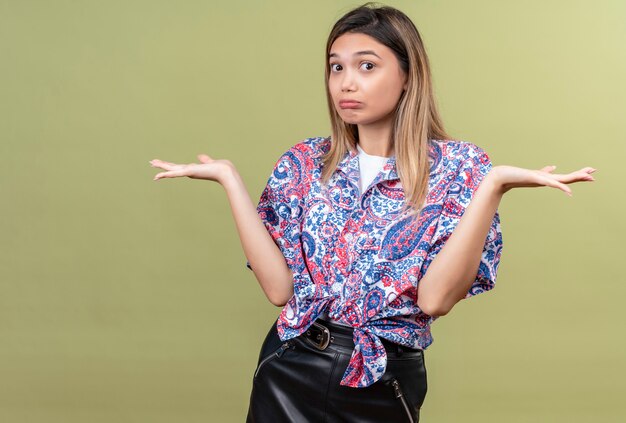 A beautiful young woman wearing paisley printed shirt showing helpless gesture and spreading her hands while looking on a green wall