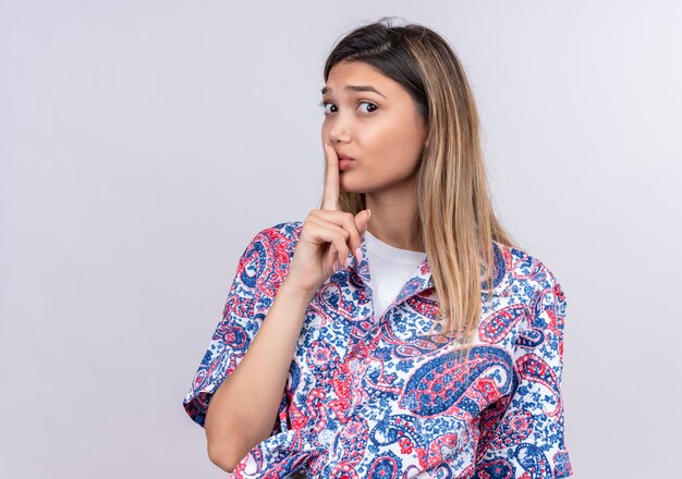 A beautiful young woman wearing paisley printed shirt showing shh gesture with finger while looking on a white wall