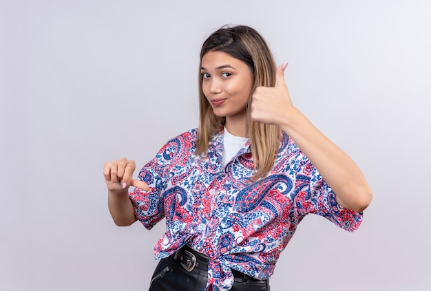 A beautiful young woman wearing paisley printed shirt showing thumbs up and down while looking on a white wall
