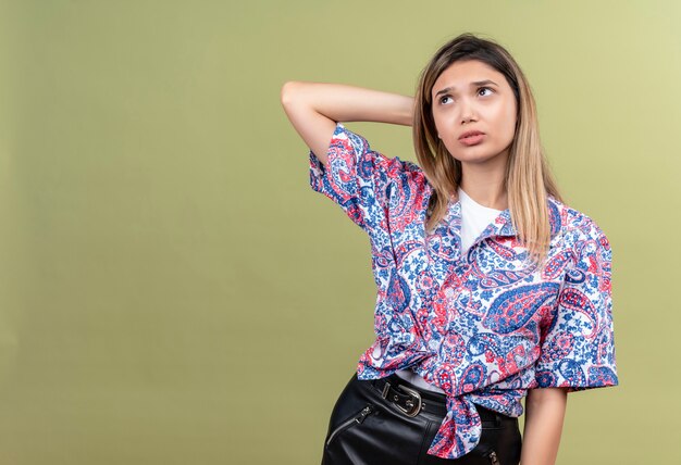 A beautiful young woman wearing paisley printed shirt thinking and holding hand on head while looking up on a green wall