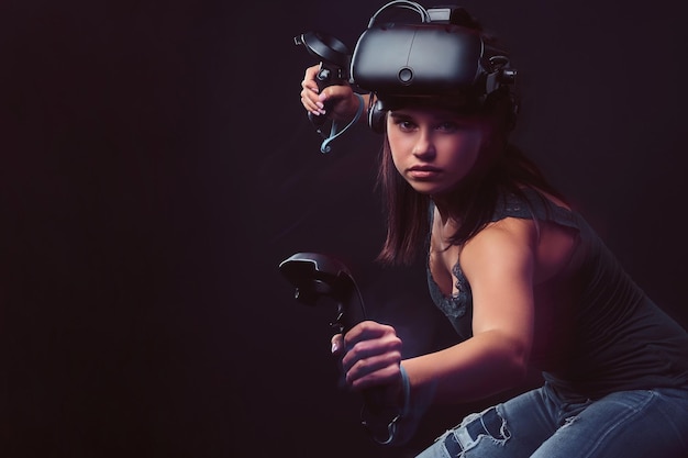 Beautiful young woman wearing virtual reality headset and holds joysticks, posing ar camera. Isolated on dark background.