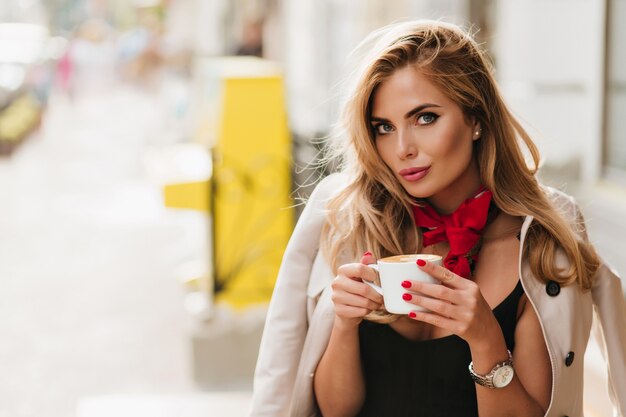 Beautiful young woman wears trendy wristwatch posing on blur background while drinks coffee after hard day