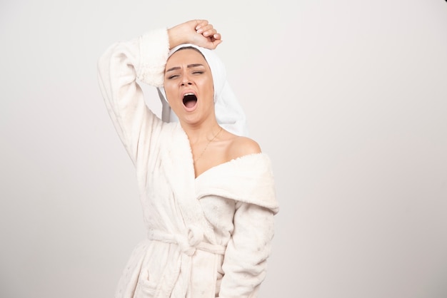 Beautiful young woman in a white bathrobe yawning on a white wall