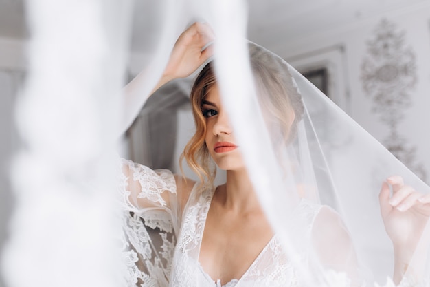 Beautiful young woman in white lingerie poses in white silk bathrobe in bright hotel room