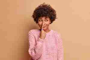 Free photo beautiful young woman with afro hair points at nose with index finger has glad expression foolishes around and looks away dressed in knitted sweater isolated over brown wall