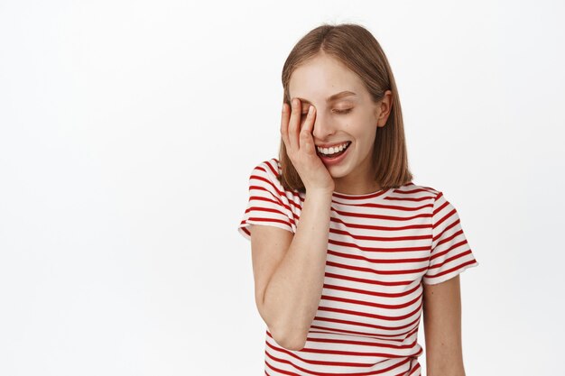 Beautiful young woman with clean, natural skin without makeup, touch her face, laughing and smiling happy with closed eyes, standing in relaxed pose against white wall.