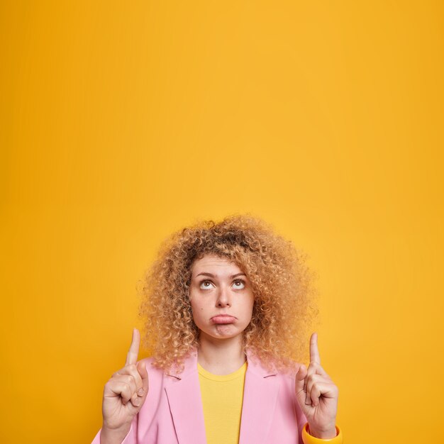 Beautiful young woman with curly bushy hair has upset face sulks and looks above shows your logo banner dressed in formal clothes isolated over yellow wall talks about something unfair