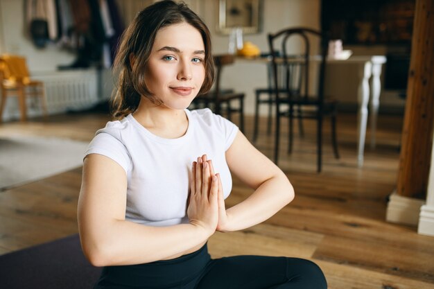 Beautiful young woman with curvy body and blue eyes sitting on floor holding hands pressed together, having refreshed relaxed facial expression after meditation