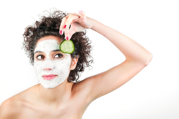 Beautiful young woman with face mask holding cucumber slice against white background