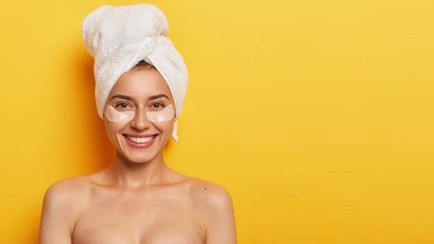 beautiful young woman with healthy delicate fresh skin under eyes, wears towel on head, applies patches for reducing fine lines, stands naked against yellow wall. Spa treatment