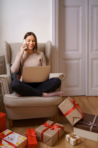 Beautiful young woman with laptop talking on mobile phone while sitting in comfortable armchair