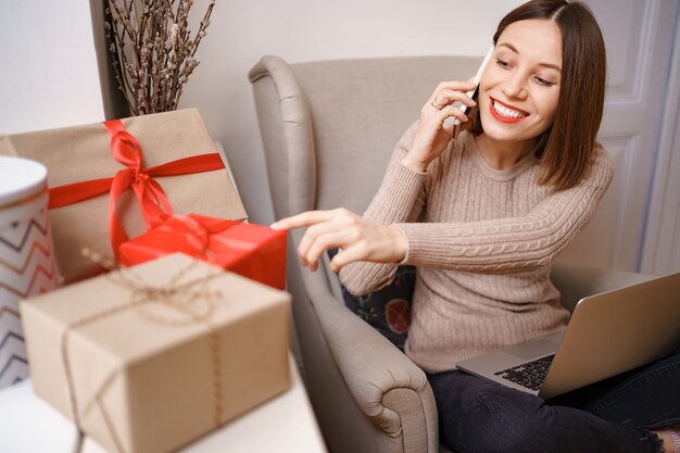 Beautiful young woman with laptop talking on mobile phone while sitting in comfortable armchair