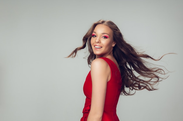 Beautiful young woman with long healthy curly hair and bright make up wearing red dress isolated on grey