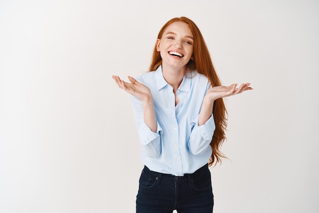 Beautiful young woman with natural red hair laughing, spread hands sideways clueless, standing over white wall