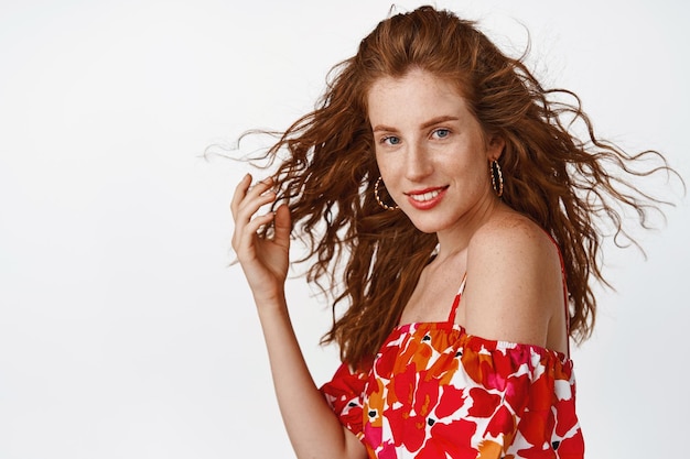 Beautiful young woman with red natural hair flying in air wind blowing at hairstyle smiling and gazing coquettish at camera standing in dress against white background