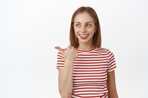 Beautiful young woman with short blond hair, pointing and looking left, reading promo text with pleased happy smile, standing in striped t-shirt over white background.