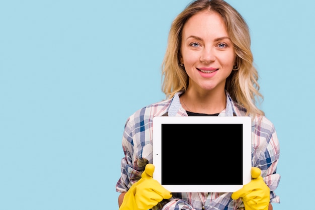 Beautiful young woman with yellow glove holding digital tablet standing against blue background