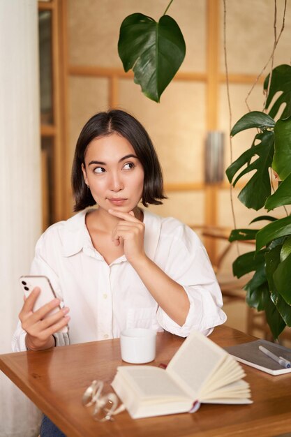 Beautiful young woman years old thinking holding smartphone and looking thoughtful sitting in cafe d