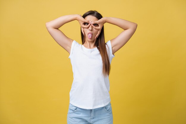 Beautiful young woman over yellow background happy face smiling doing ok or glass sign with hand on eye looking through fingers.