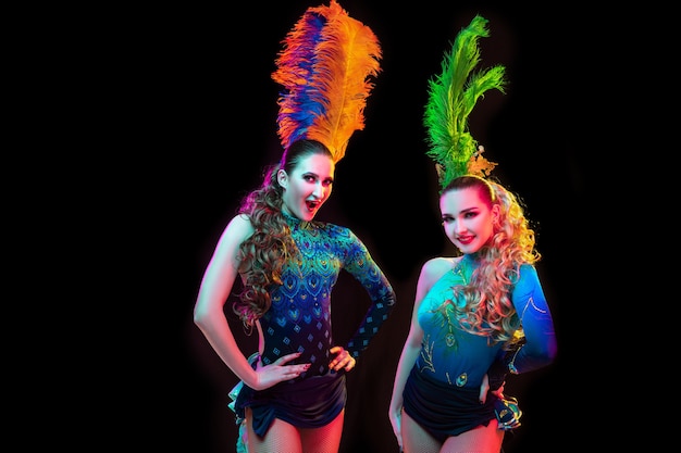 Beautiful young women in carnival, stylish masquerade costume with feathers on black background in neon light.
